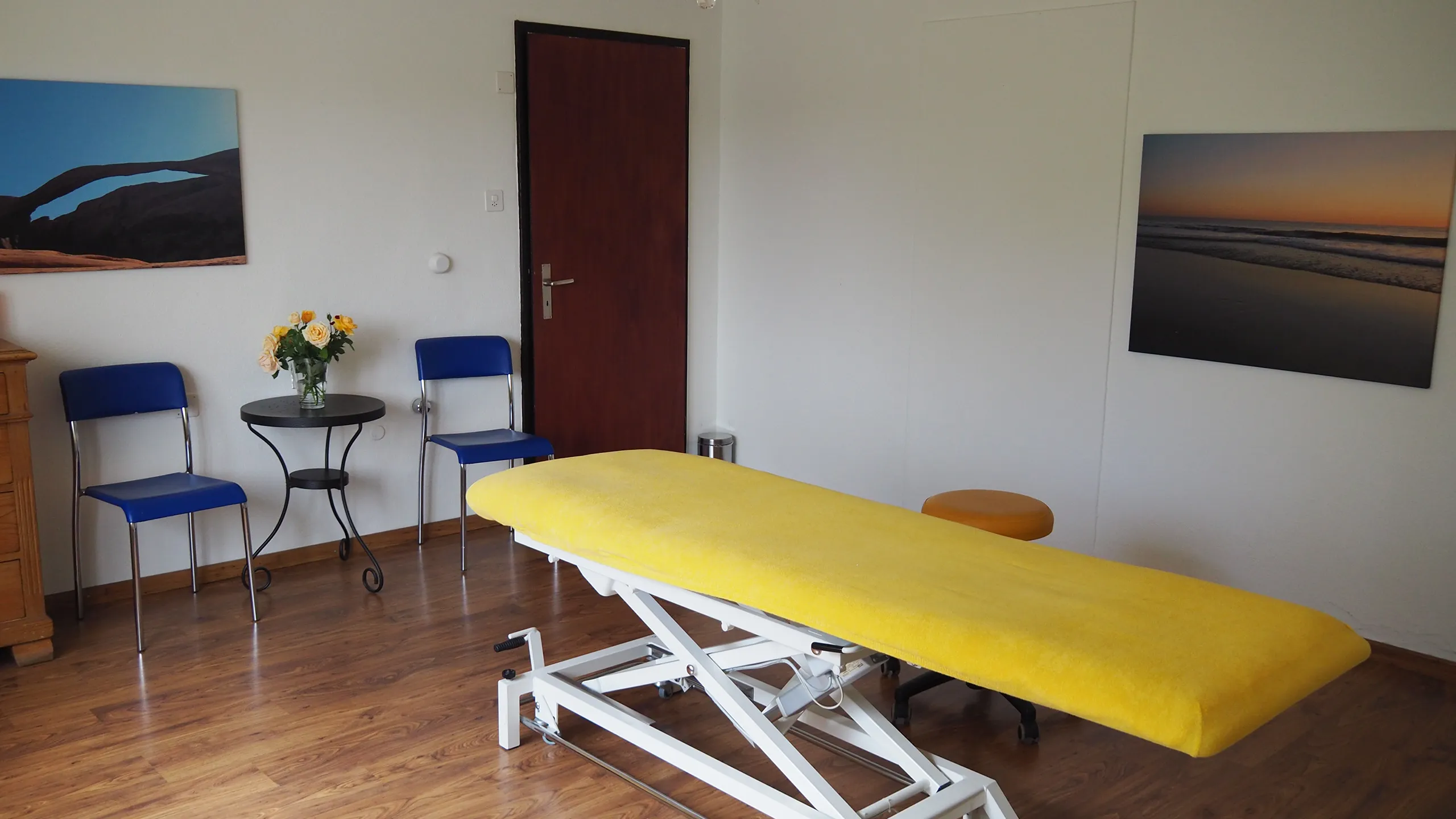Der Behandlungsraum der Praxis mit Therapieliege, Wartestühlen und Blumen